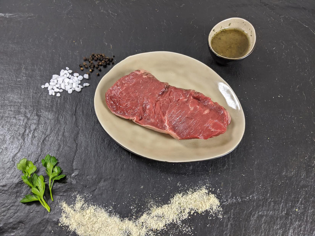 Rumpsteak Pfeffer und Salz auf Teller angerichtet mit einer Schale Marinade groben Salz und Pfefferkörnern und Kräutern auf Schieferhintergrund
