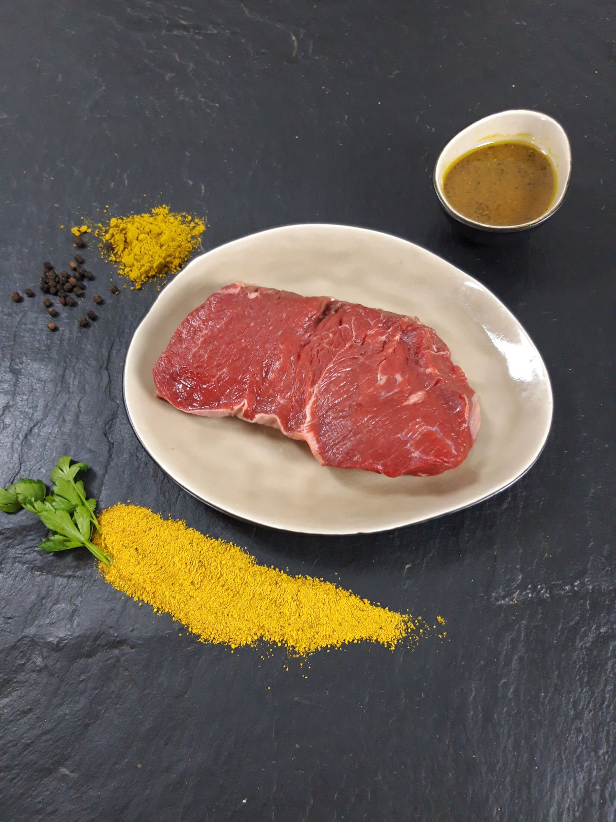 Rumpsteak Bombay auf Teller angerichtet mit einer Schale Marinade Kräutern und Currypulver auf Schieferhintergrund