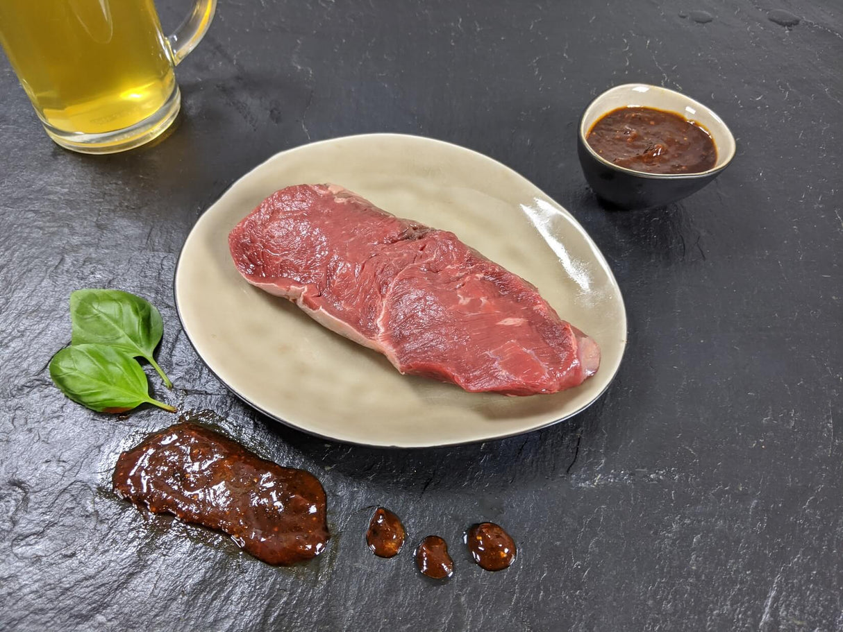 Rumpsteak Braumeister auf Teller angerichtet mit einer Schale Marinade Kräutern und einem Glas Bier auf Schieferhintergrund