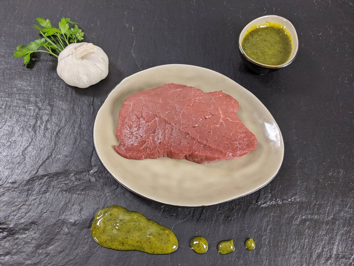 Rinderhüftsteak Kräuter Knoblauch auf Teller angerichtet mit einer Schale Marinade Kräutern und einer Knoblauchknolle auf Schieferhintergrund