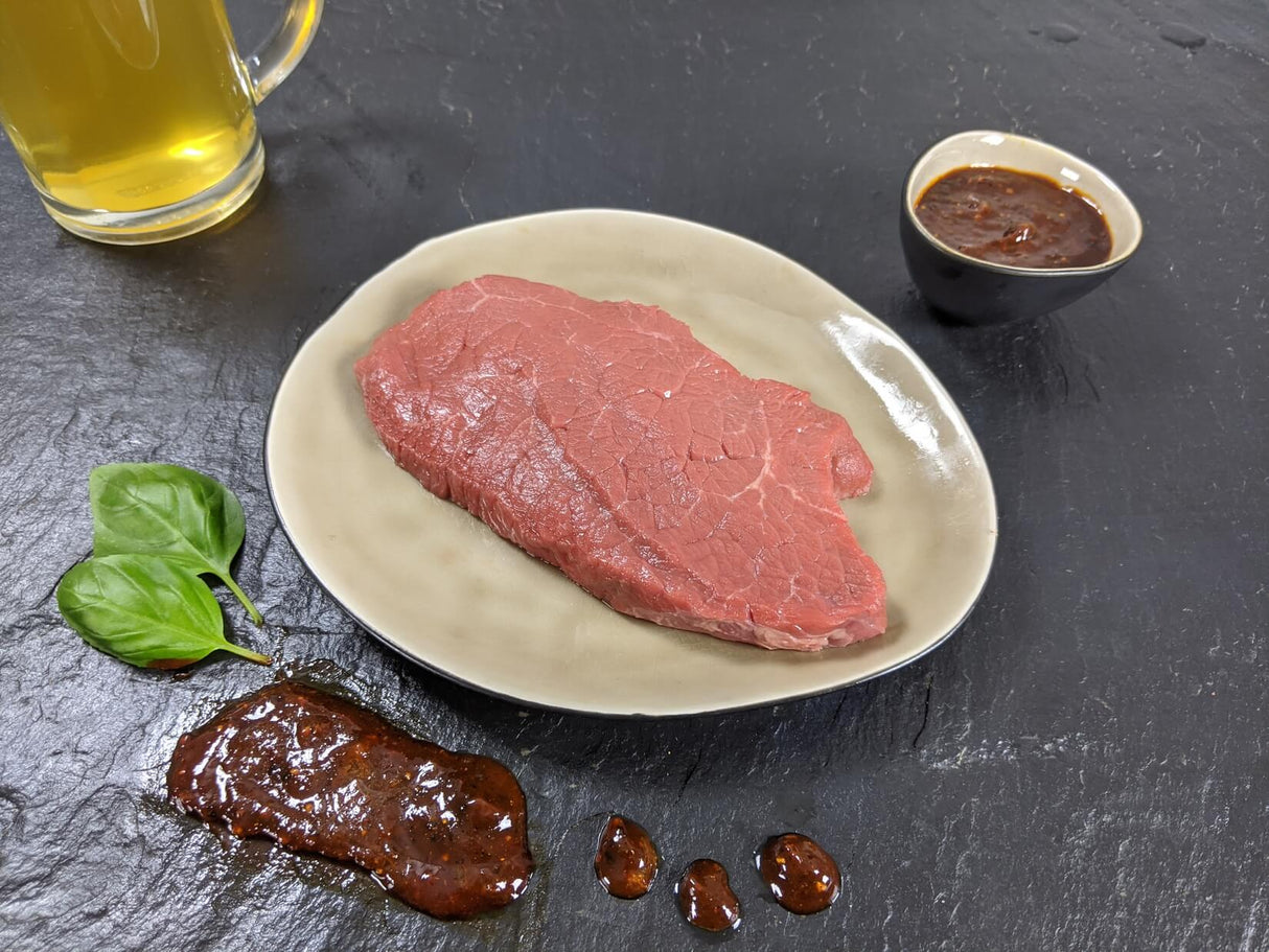 Rinderhüftsteak Braumeister auf Teller angerichtet mit Schale Marinade Kräutern und einem Glas Bier auf Schieferhintergrund