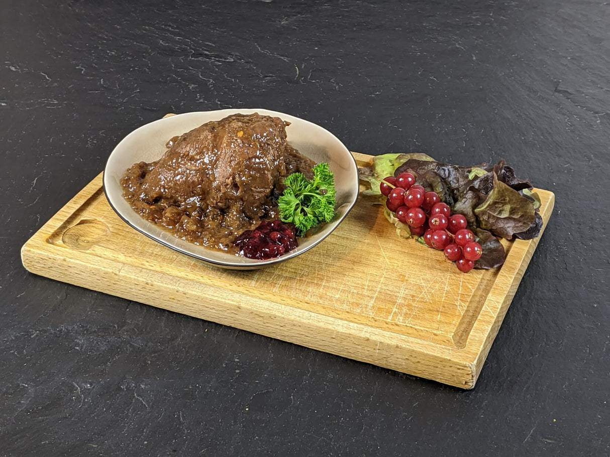 Rinderbäckchen gegart in Sauce im Teller angerichtet mit Preiselbeeren Kräutern und Salat auf Holbrett und Schieferhintergrund