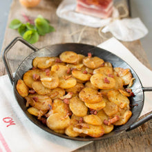 Bratkartoffeln gegart auf Holzbrett mit Serviertte und Speck Basilikum und Zwiebeln im Hintergrund