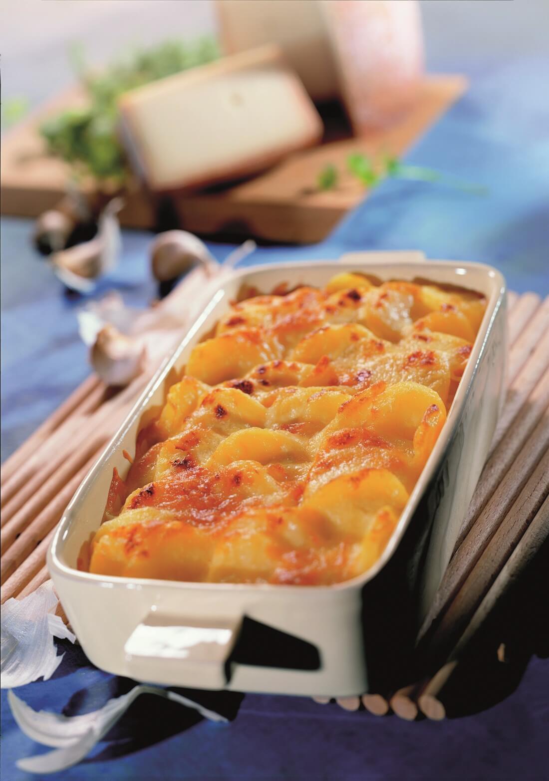 Kartoffelgratin auf Bambusteller in Auflaufform mit Käse im Hintergrund