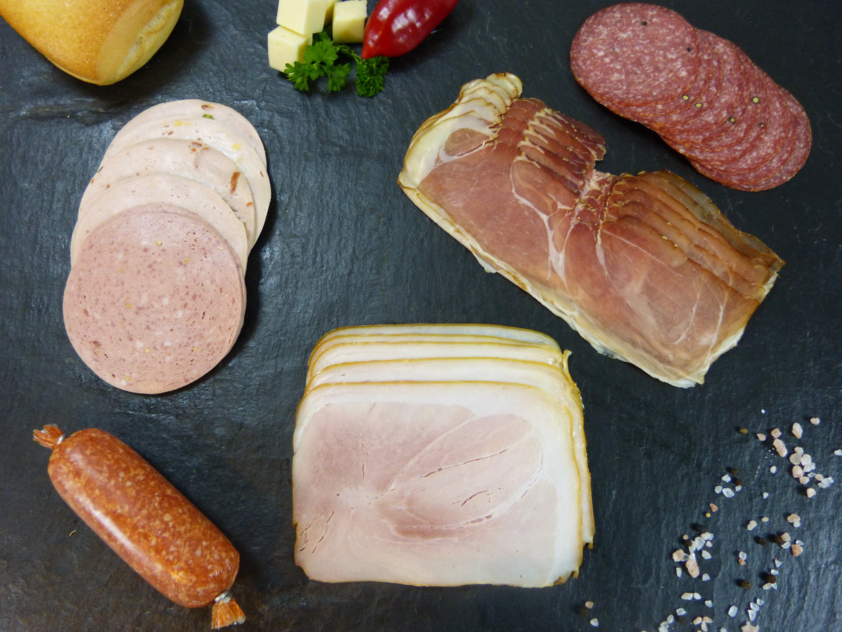 Auswahl aus Bauernschinken und Metzgerschinken vom EIFEL Schwein Brühwurstaufschnitt gemischt Pfeffersalami und Zwiebelmettwurst angerichtet auf Schieferhintergrund mit Käsewürfeln, Chilischoten und einem Brötchen