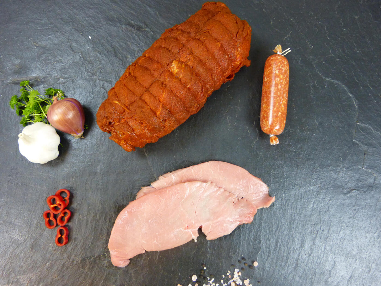 Auswahl aus Kammspießbraten handgerollt Kalb Oberschalenschnitzel und Zwiebelmettwurst auf Schieferhintergrund angerichtet mit Chili Knoblauchknolle Zwiebel und Petersilie