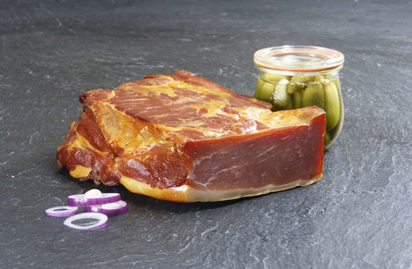 Schinkenspeck angerichtet mit eingelegten Gurken und roten Zwiebelringen auf Schieferhintergrund