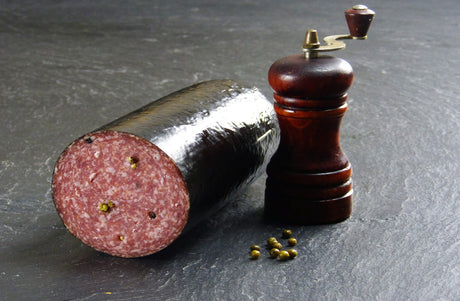 Pfeffersalami am Stück angerichtet mit Pfeffer und einer Pfeffermühle auf Schieferhintergrund