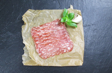 Salami Spianata Romana in scheiben geschnitten angerichtet mit Basilikum und einer Knoblauchzähe auf Backpapier mit Schieferhintergrund