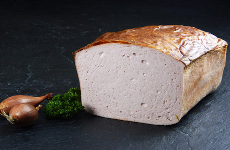 Ein halber Fleischkäse gebacken vom EIFEL Schwein angerichtet mit zwei kleinen roten Zwiebeln und Petersilie auf Schieferhintergrund