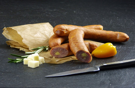 Zwei Käsebrater vom EIFEL Schwein un ein halbierter Käsebrater angerichtet mit Käsewürfeln Thymian und einer gelben Paprika mit einem Messer an der Seite auf Backpapier mit Schieferhintergrund