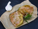 Schweinerückensteak mit Kräuter Knoblauch marinert auf Backpapier dekoriert mit einer Knoblauchknolle und Kräutern auf Schieferhintergrund