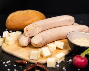 Angeschnittene Bratwurst Chili-Cheese auf einem Holzbrett, dekoriert mit Käsewürfeln, Chilischoten und Gewürzen. Außerdem ein Radieschen und Schale mit Dip sowie einem Brötchen im Hintergrund