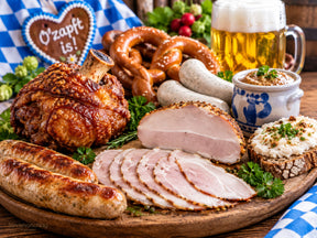 Haxe, Krustenbraten, Käsebratwürste, Schmalzbrot, Brezel, Bierkrug, lecker angerichtet auf einer Holzplatte mit Bayrischem Tuch im Hintergrund und einem Lebkuchenherz mit der Aufschrift o´zapft is!