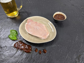 Schweinerückensteak Braumeister auf Teller angerichtet mit einer Schale Braumeister Marinade Kräutern und einem Glas Bier auf Schieferhintergrund