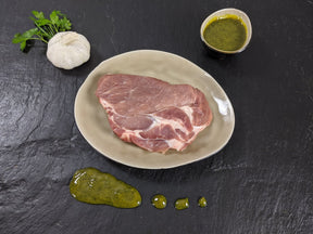 Schweinebauchscheibe Kräuter Knoblauch auf Teller angerichtet mit einer Schale Marinade Knoblauchknolle und Kräutern auf Schieferhintergrund