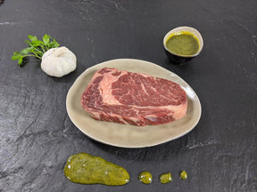 Entrecôte Kräuter Knoblauch auf Teller angerichtet mit einer Schale Marinade Knoblauchknolle und Kräutern auf Schieferhintergrund