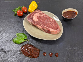 Entrecôte Barbecue auf Teller angerichtet mit einer Schale Marinade Tomaten Paprika und Kräutern auf Schieferhintergrund