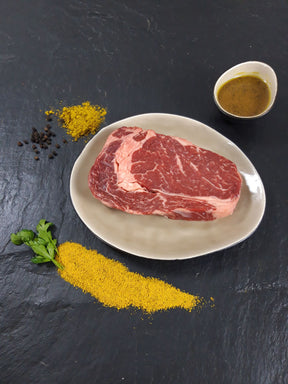 Entrecôte Bombay auf Teller angerichtet mit einer Schale Marinade Kräutern und Currypulver auf Schieferhintergrund