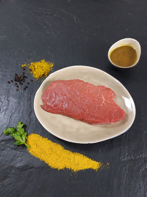 Rinderhüftsteak Bombay auf Teller angerichtet mit Schale Marinade Kräutern und Currypulver auf Schieferhintergrund